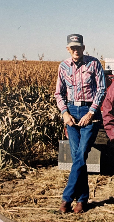 A younger Grant Fletcher leans against a truck wearing blue jeans, a collared long-sleeve shirt and a ball cap next to a corn field.