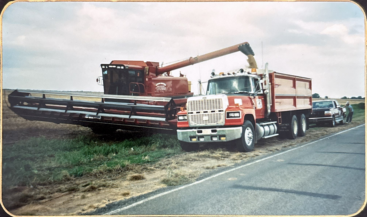 A combine augers grain into the box of a tandem grain truck next to a paved roadway in the 80s or 90s.