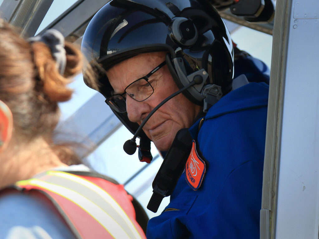 An older man sits in the pilot seat of a helicopter wearing a helmet with a radio mike boom attached.