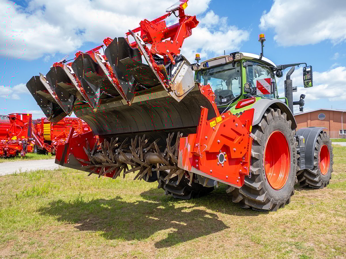 A tractor is parked with its rear-mounted tiller lifted up into the air showing the tiller's blades and the axle they are mounted on.