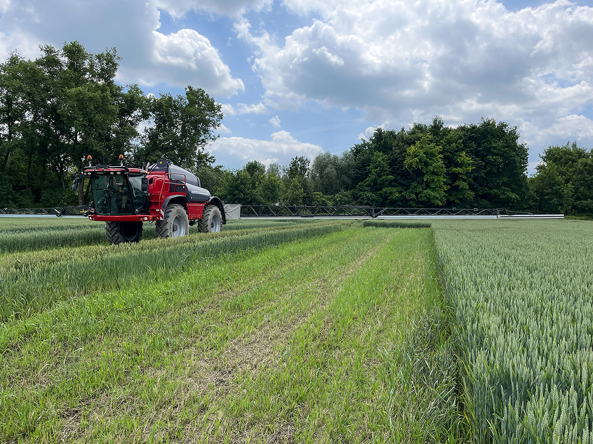 A Horsch sprayer with a very wide boom moves over a crop on a sunny day with scattered clouds.