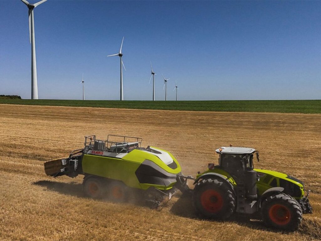 A promotional drone photo of the Class 70 t sqare baler in action in a field beneath a number of large wind turbines.