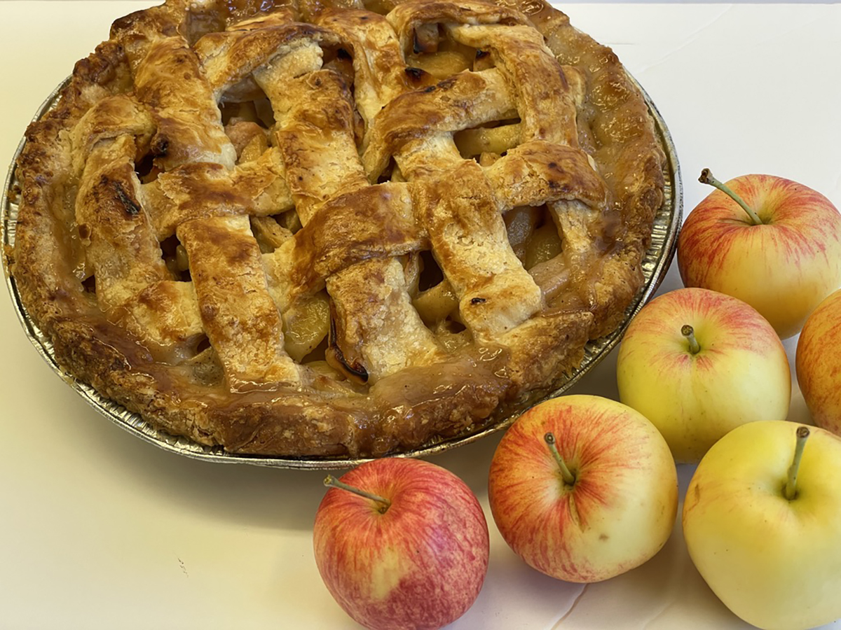 A freshly-backed apple pie with a lattice-like crust on top sits on a white surface next to a few apples.