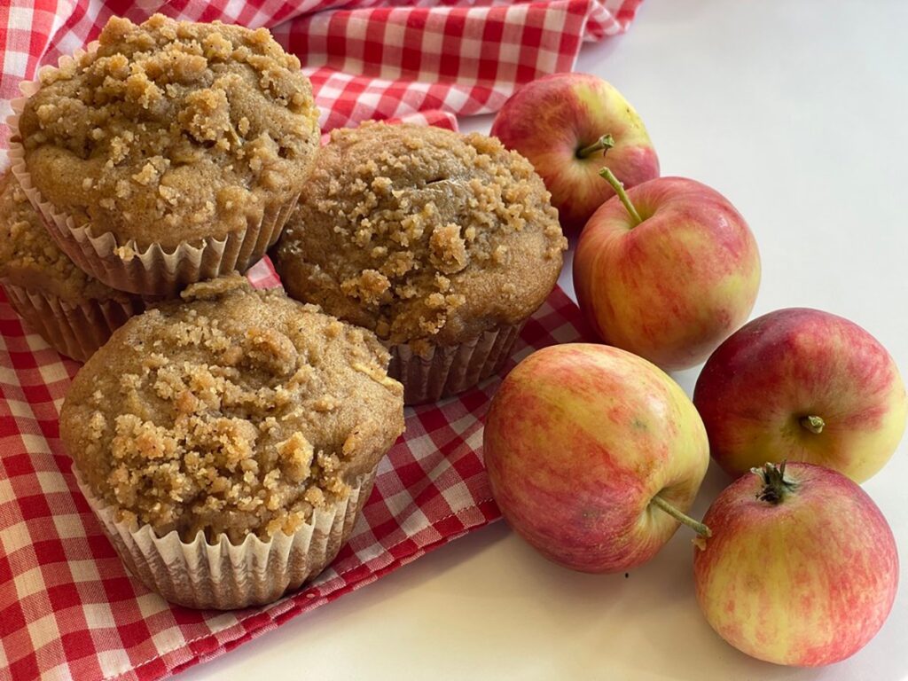 Several freshly-baked muffins sit next to a few apples on a red and white checkered tablecloth.