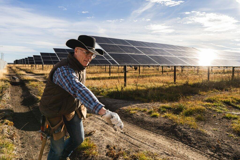 A man in a black cowboy hat wearing work gloves and a vest with a tool belt over his blue jeans stands in front of a large solar array.