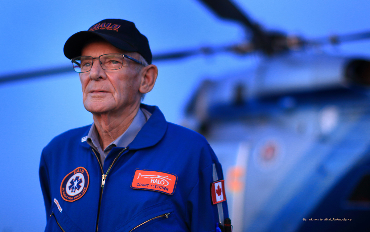 Grant Fletcher poses for a portrait in his HALO ball cap and blue flight suit with a helicopter blurred in the background.