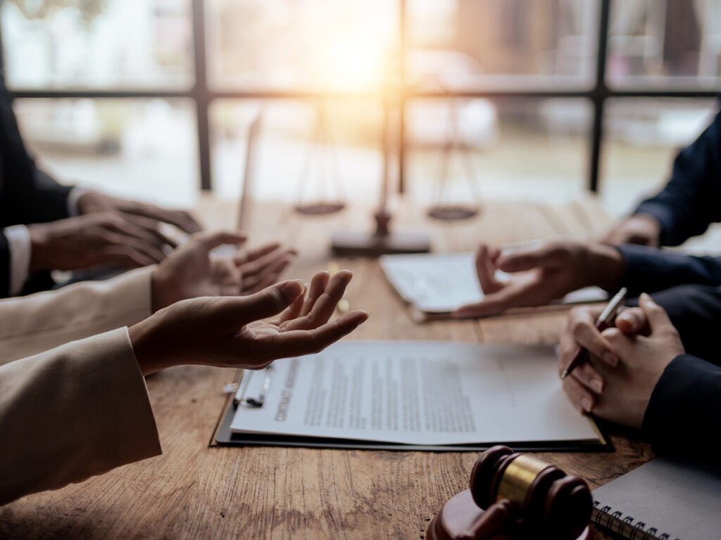 The animated hands of people seated on opposite sides of a boardroom table with a gavel in the foreground, and a blurry 