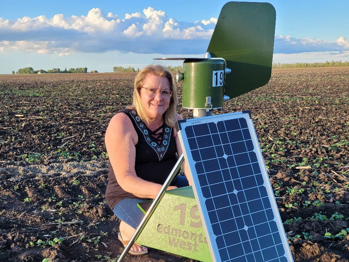 A woman kneels in a potato field next to a round, green metal cylinder - a spore trap - mounted on a tripod that has a solar panel on it.