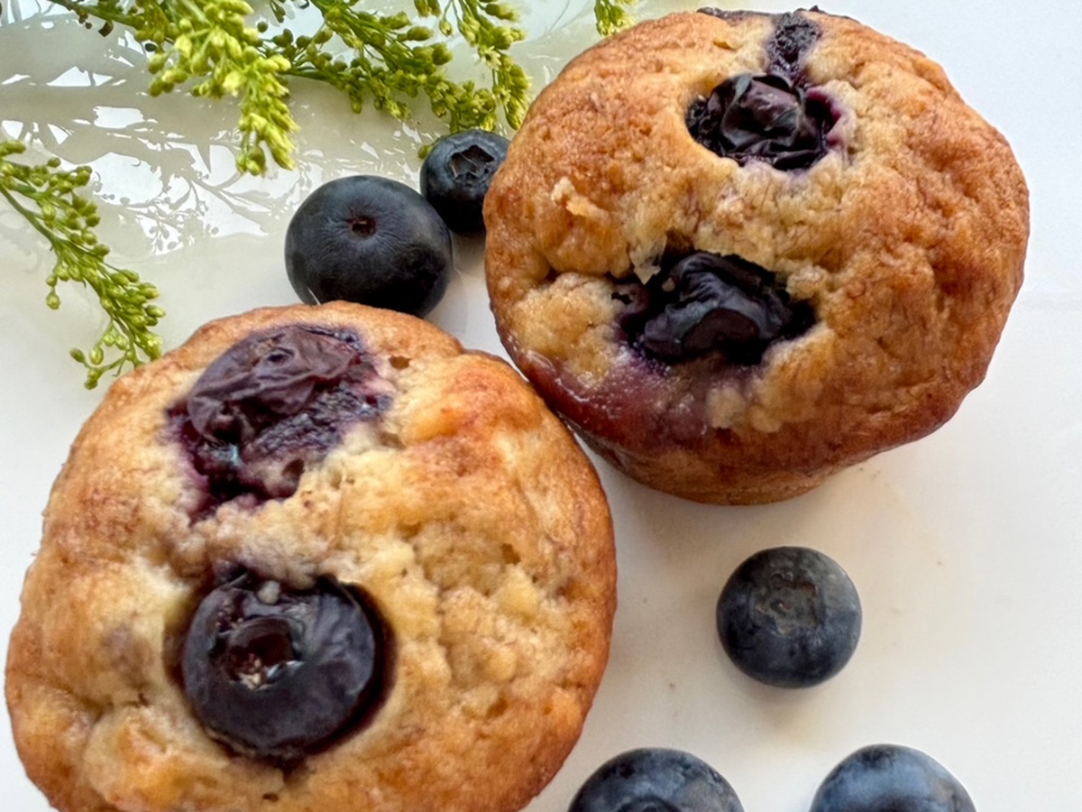 Two banana blueberry muffins on a white surface with a few blueberries beside them.