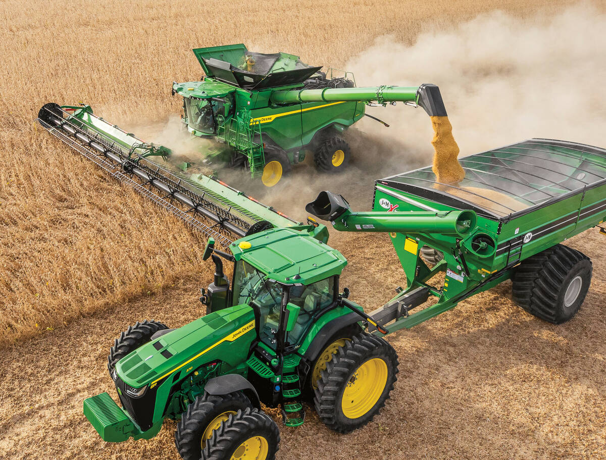 A stock photo provided by John Deere showing, from above, a combine harvesting corn and augering it into a grain cart being pulled by a tractor beside it.