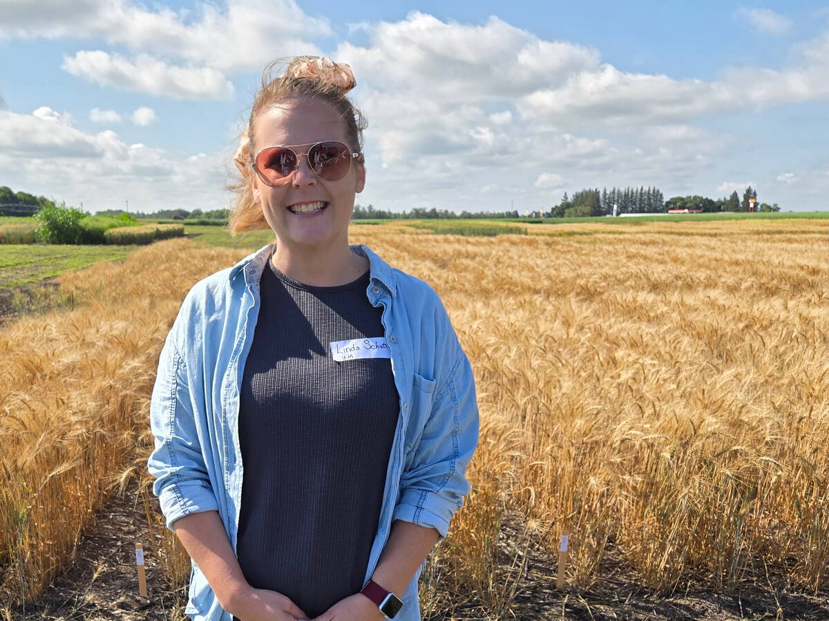 A woman in a light blue denim shirt with a dark blue shirt underneath poses for a photo, smiling, in front of a wheat field.