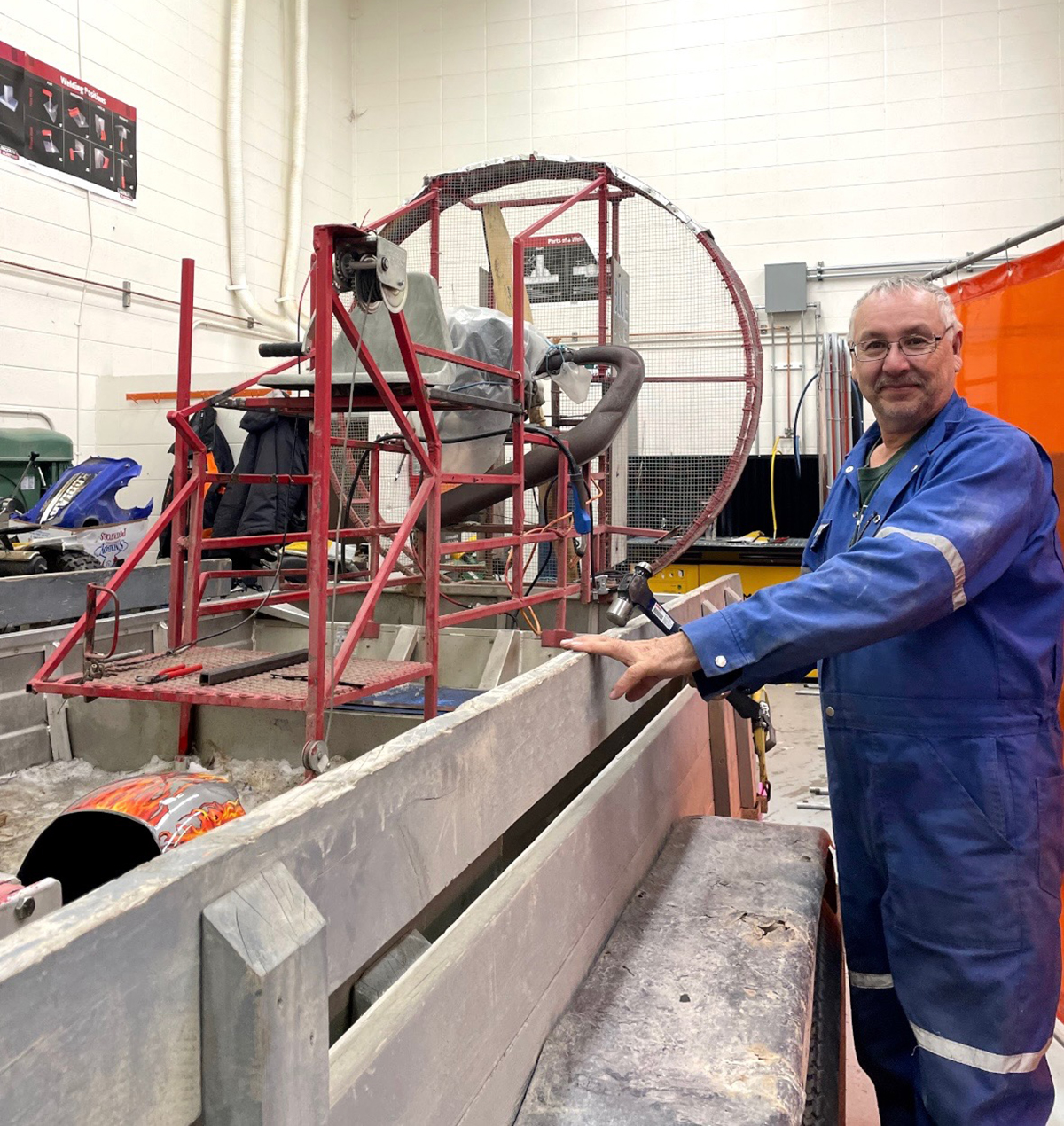 A man in blue coveralls stands in a shop beside an air boat that serves as a wild rice harvester.