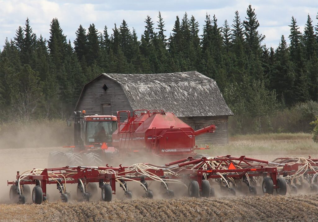 A tractor kicks up dust as it pulls a seeder planting winter wheat with an old barn and some pine trees in the background.