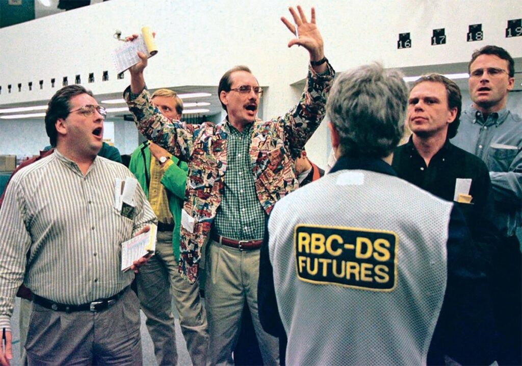 A trader on the floor of the Winnipeg Commodity Exchange stands, clad in a jacket with a busy pattern, hands raised above his head with a pen in one hand and a receipt-like book in the other, as other traders stand around him.