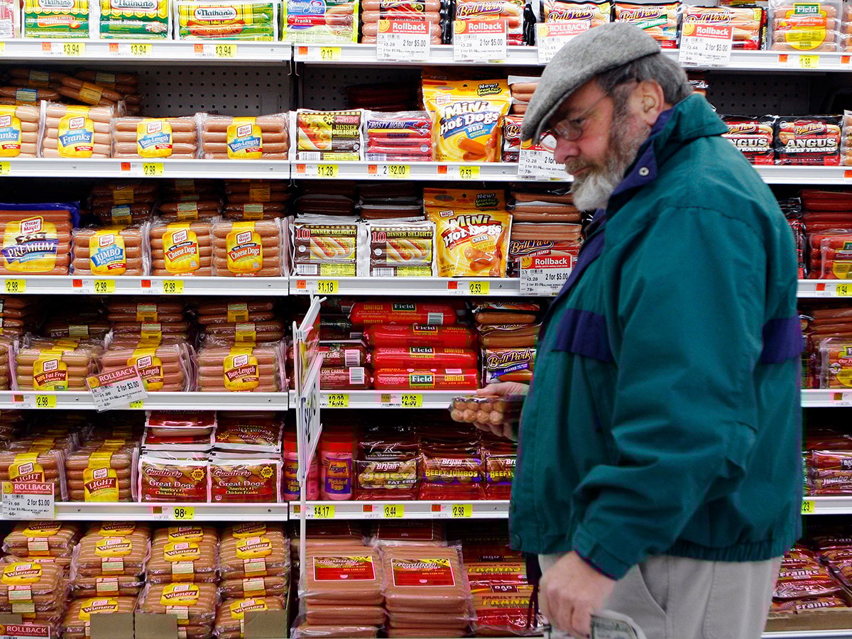 A bearded middle-aged man wearing glasses and a tam-style hat picks up a package of wieners in a WalMart grocery store.