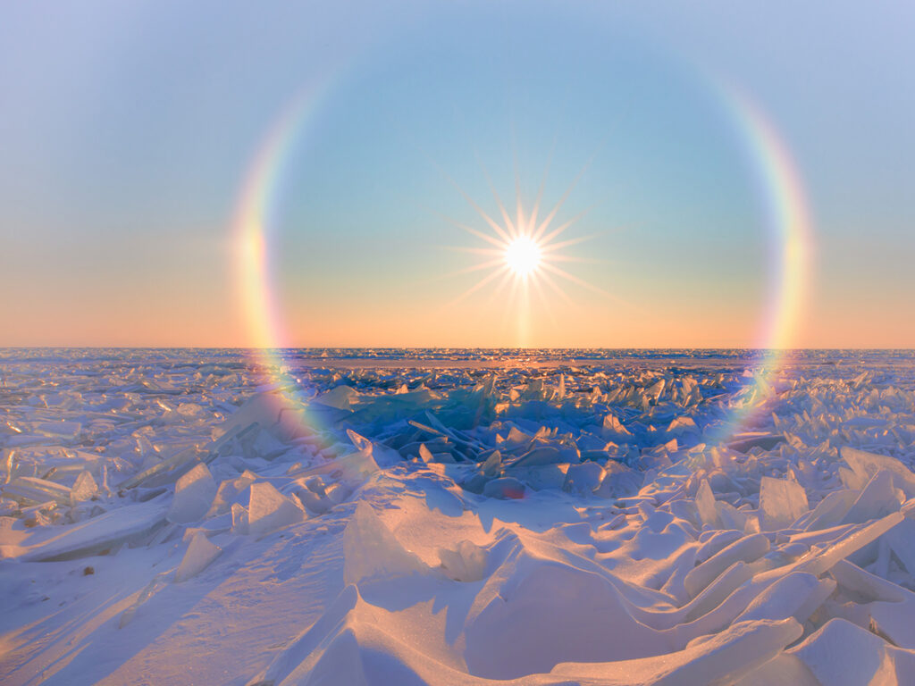 Ice crystals form a halo around the sun that sits low in the sky over a frozen, snowscape below.