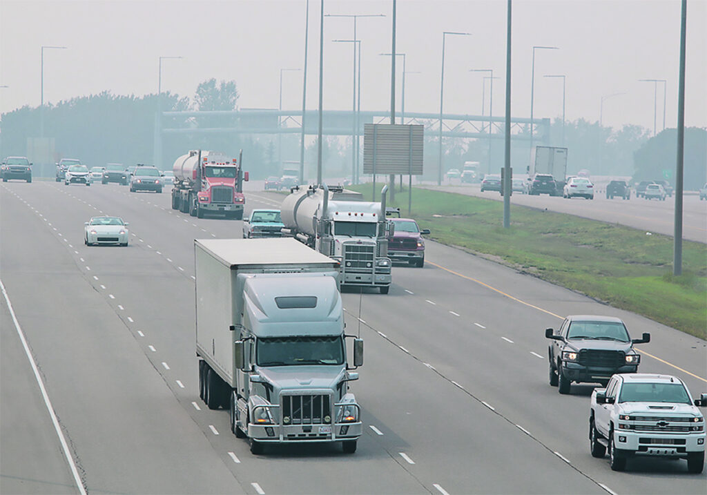 A number of large tractor trailer units scattered among the traffic on a busy divided highway.