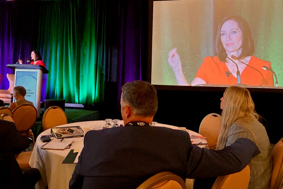 Two people seated at a round table near the stage at a conference look toward a woman speaking at a podium as her image is simultaneously projected onto a large screen behind her for those further away to see.