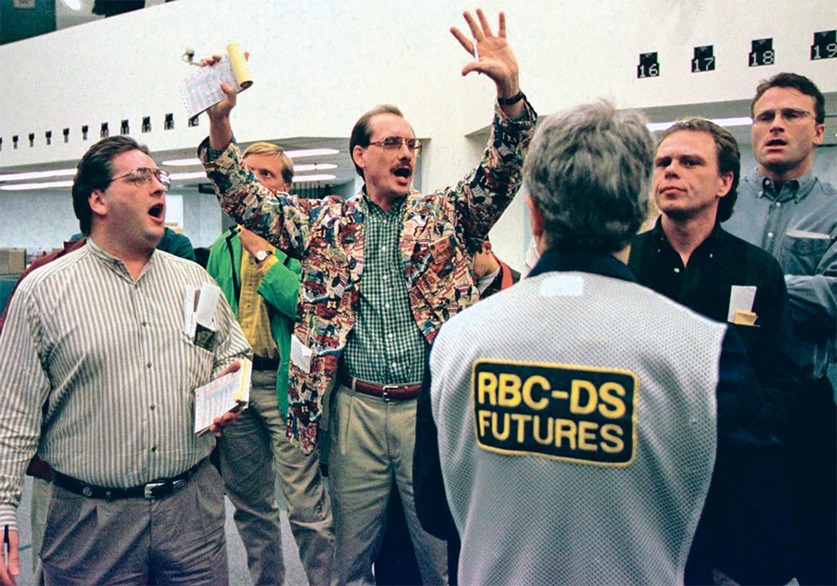 A trader on the floor of the Winnipeg Commodity Exchange stands, clad in a jacket with a busy pattern, hands raised above his head with a pen in one hand and a receipt-like book in the other, as other traders stand around him.