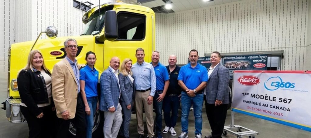 A group of men and women stand in front of a large yellow truck next to a sign with a red, oval Peterbilt logo on it that reads (in french) 