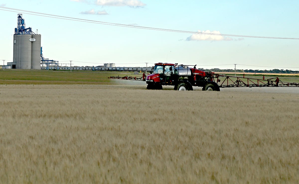 A high clearance sprayer sprays a crop with a concrete inland grain terminal visible in the background.