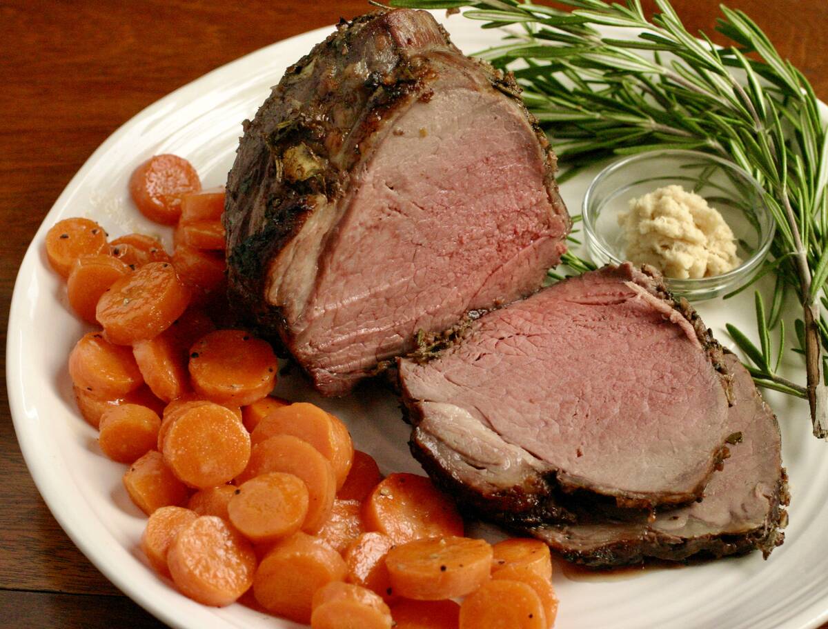 A large protion of roast beef with two slices cut and laying on a white dinner plate next to some glazed carrots, a small glass bowl with some horse raddish in it and a sprig of rosemary.