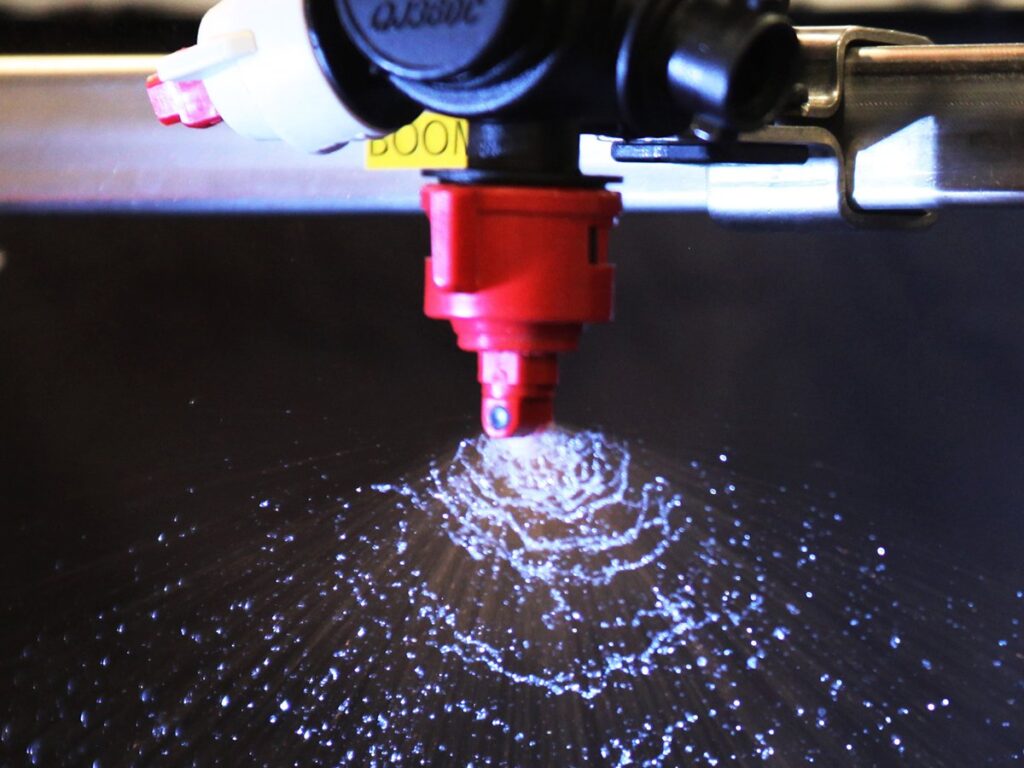 A close-up of a single nozzle on a sprayer with the spray droplets fanning out and backlit against a black background.
