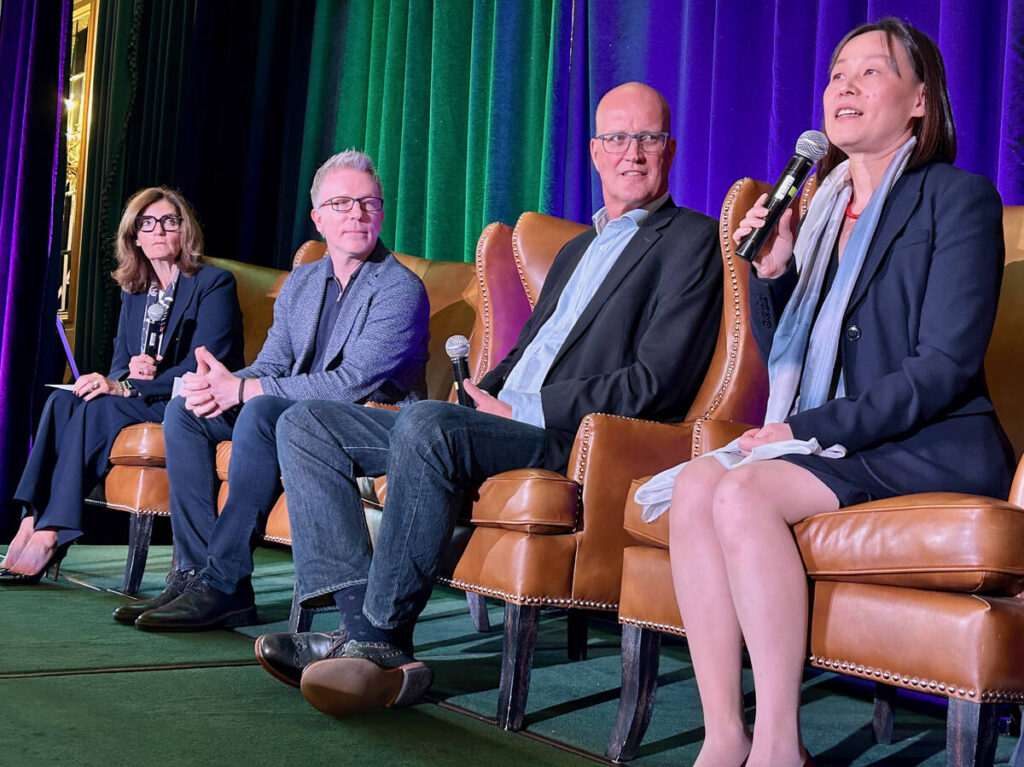 Four people, two women and two men, sit on comfortable brown leather wing-back chairs arranged in a straight line beside one another on a stage during a conference. Each has a microphone in their hands and the woman closest to the camera is speaking into it.