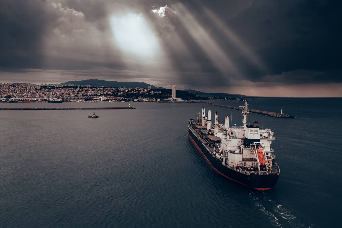 A cargo ship approaches port under a cloudy sky.