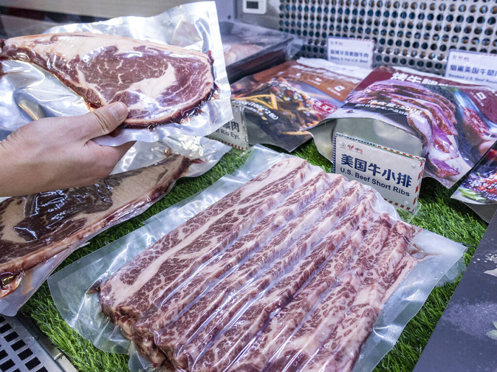 A hand holds a vacuum-packed steak above a meat display in a grocery story with a sign written in Chinese characters, subtitled in english below, 