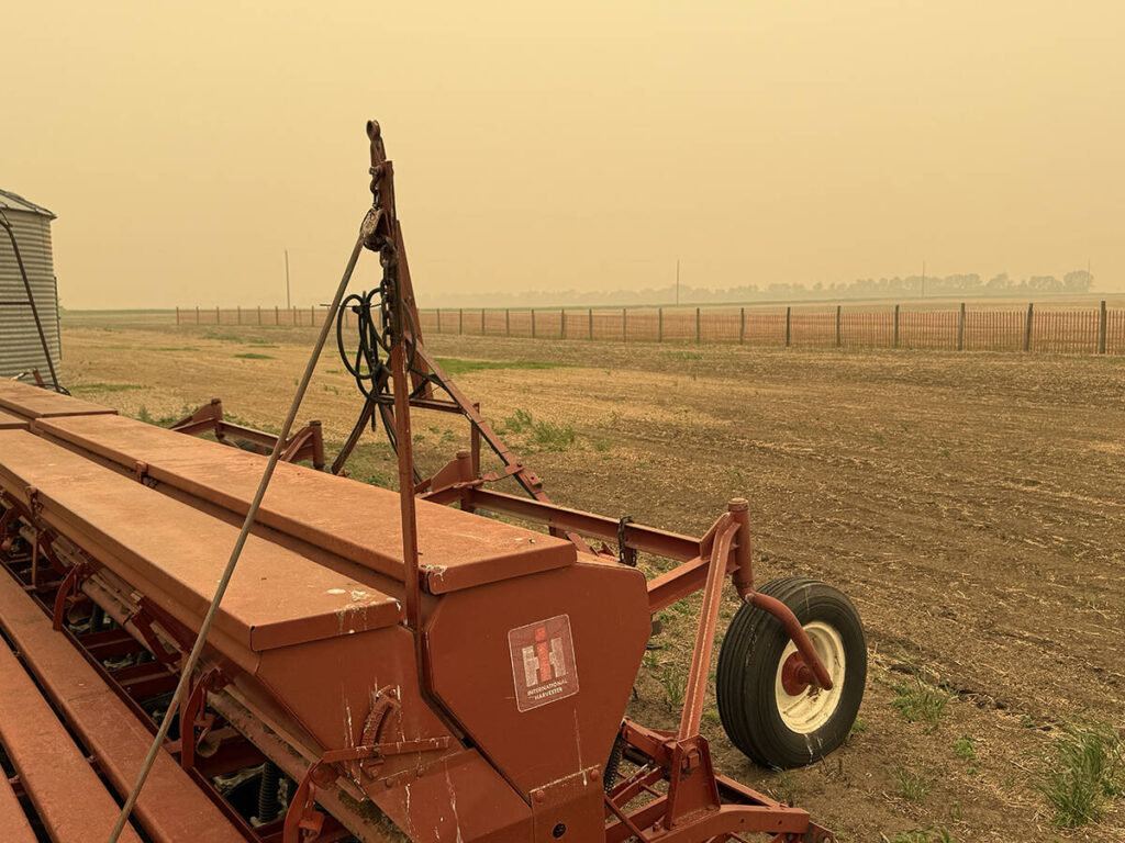 Thick smoke from northern fires can be seen in the distance while an older seeder and a steel grain bin are visible in the foreground.