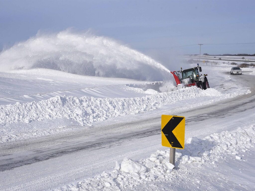 A tractor with a large, front-mounted snowblower throws snow high into the air as it clears a road.