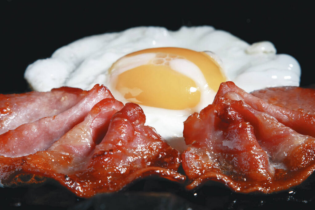 A fried egg, sunny side up, on a black surface with two pieces of crispy bacon in front of it.
