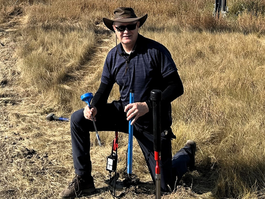 A man wearing an Aussie-style cowboy hat and sunglasses kneels in a field as he appears to pounding some sort of probe into the earth. A hammer is visible on the ground behind him.