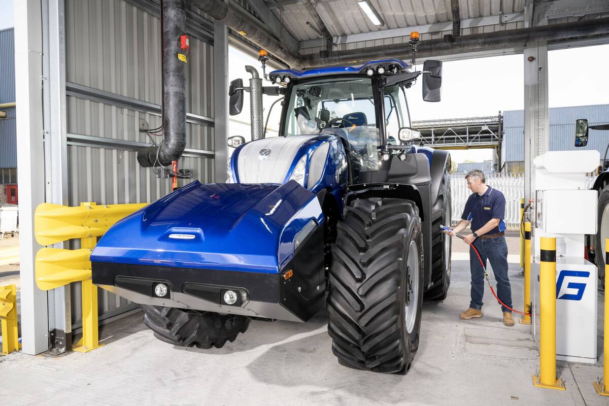 A man holding a fuel hose prepares to refuel a large, blue methane-powered tractor beneath the tin roof of an outdoor refuelling station.