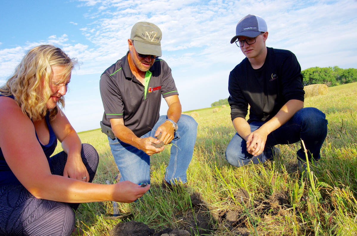 Three people, a woman on the left and two men, crouch in a farm field examining the soil that two of them hold in their right hands.