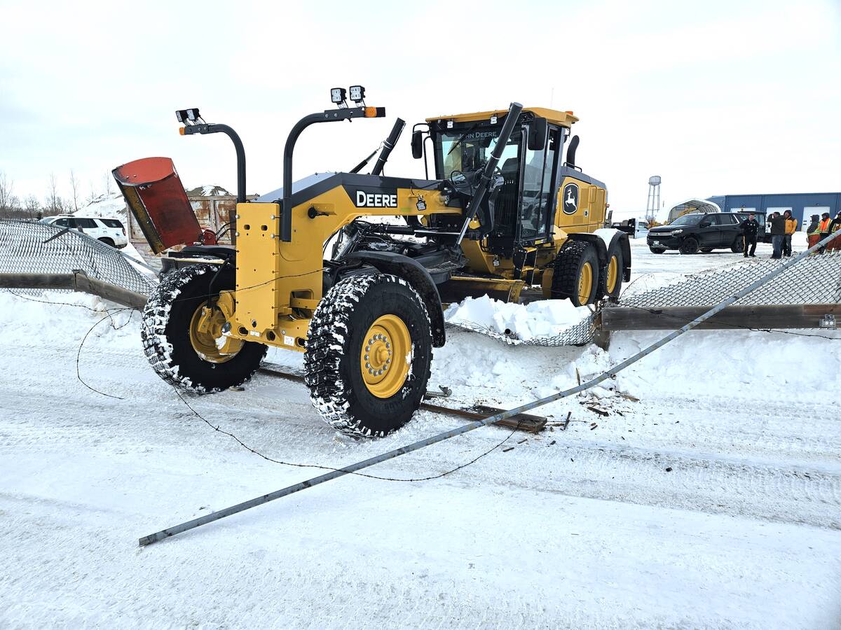 A small crowd of people gather in a snowy parking lot looking at a rod grader that has smashed through a fence after being stolen.
