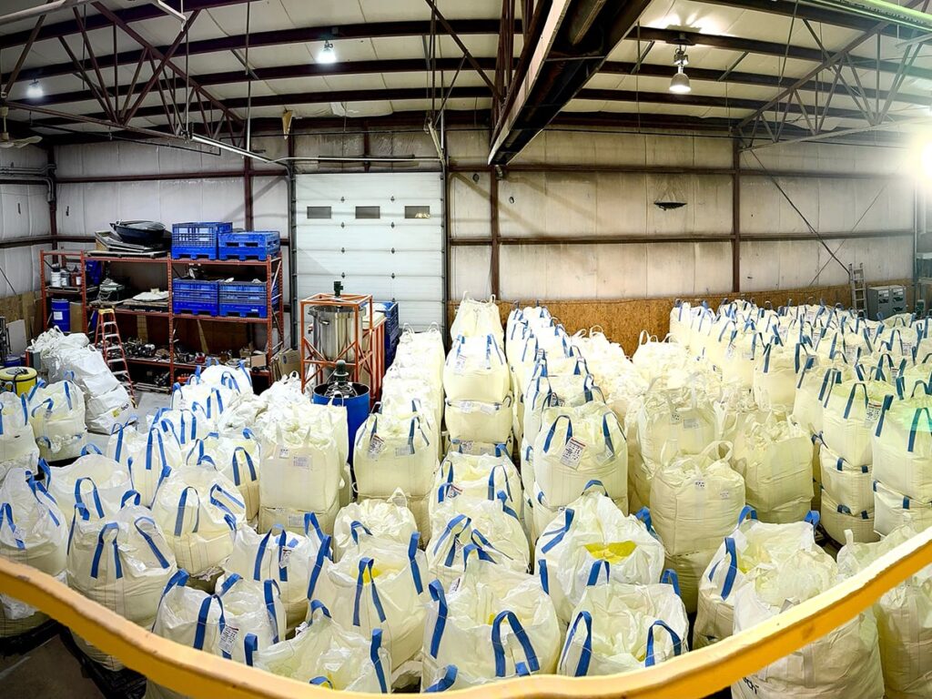 Dozens of bulging white bulk bags with blue straps on each corner sit in a warehouse ready for distribution.