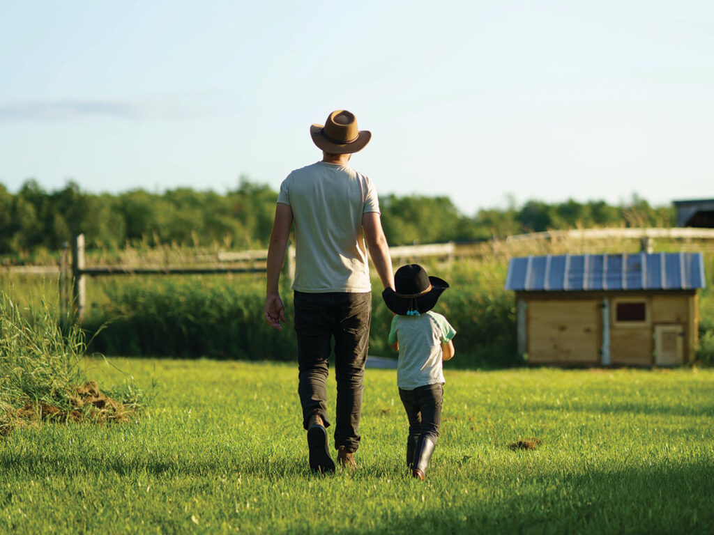 A farmer and his young son, both wearing cowboy hats, hold hands as they walk away from the camera on green grass toward a small shed in the distance.