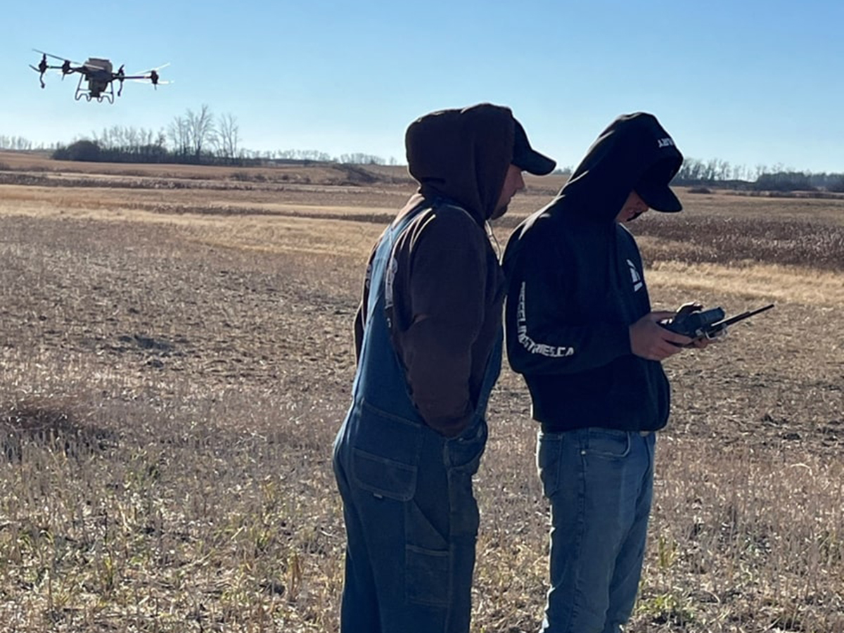 Two young men stand in a field, one holding aremote control while a quad-copter-style drone flies just above head height in the background behind them.