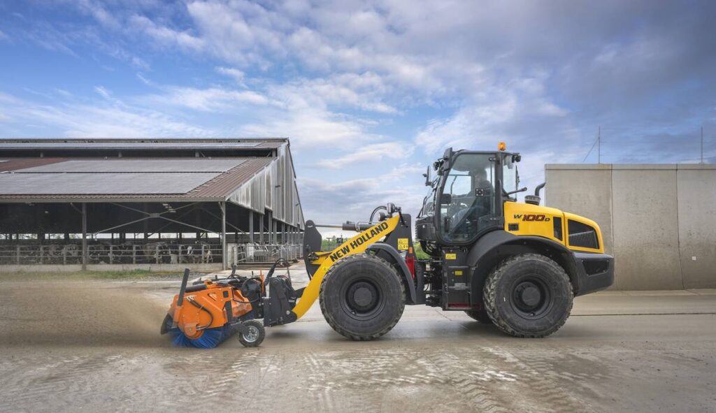 A promotional image of New Holland&rsquo;s new W100D wheel loader using a broom attachment to sweep a large concrete apron with a dairy barn and cattle visible in the background.