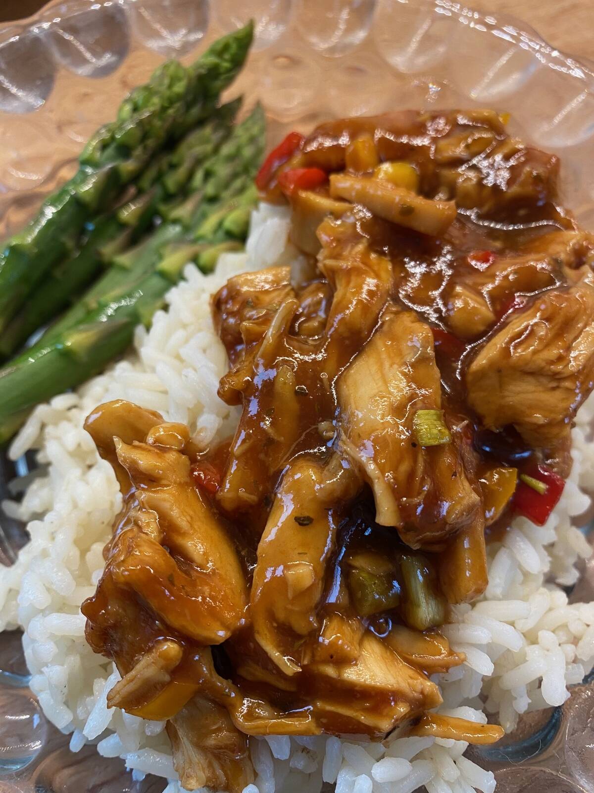A close up of some smaller pieces of chicken breast smothered in apricot sauce on a bed of white rice with some asparagus spears on the side of the plate.