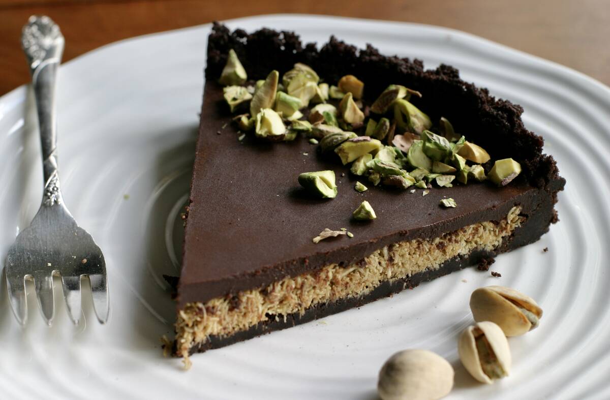 A chocolate-topped pie-like wedge with some chopped nuts on top sits on a white plate with a fork beside it.