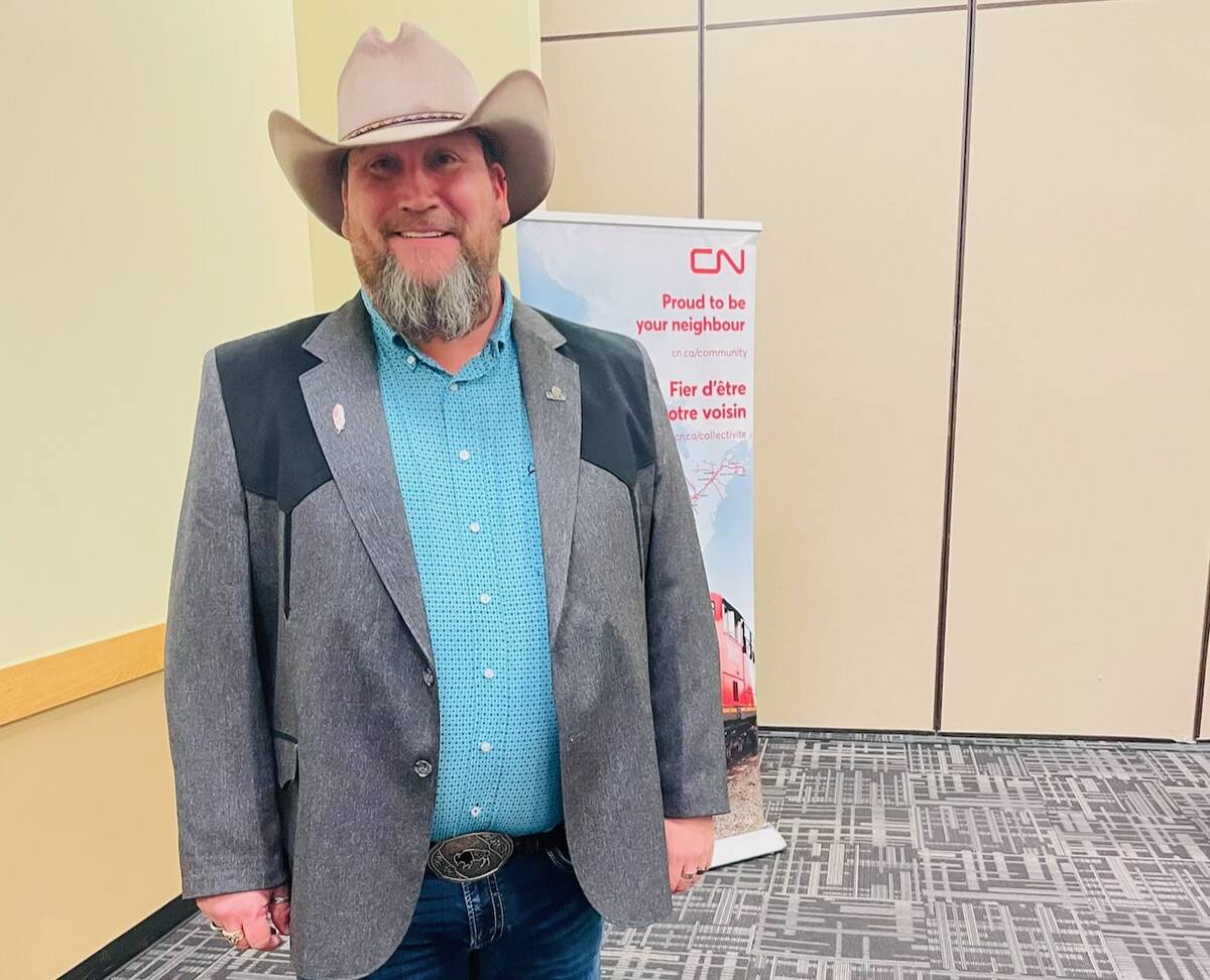 A smiling, bearded man in a cowboy hat and a western-style suit jacket wearing jeans with a large buckle on his belt poses for a photo.