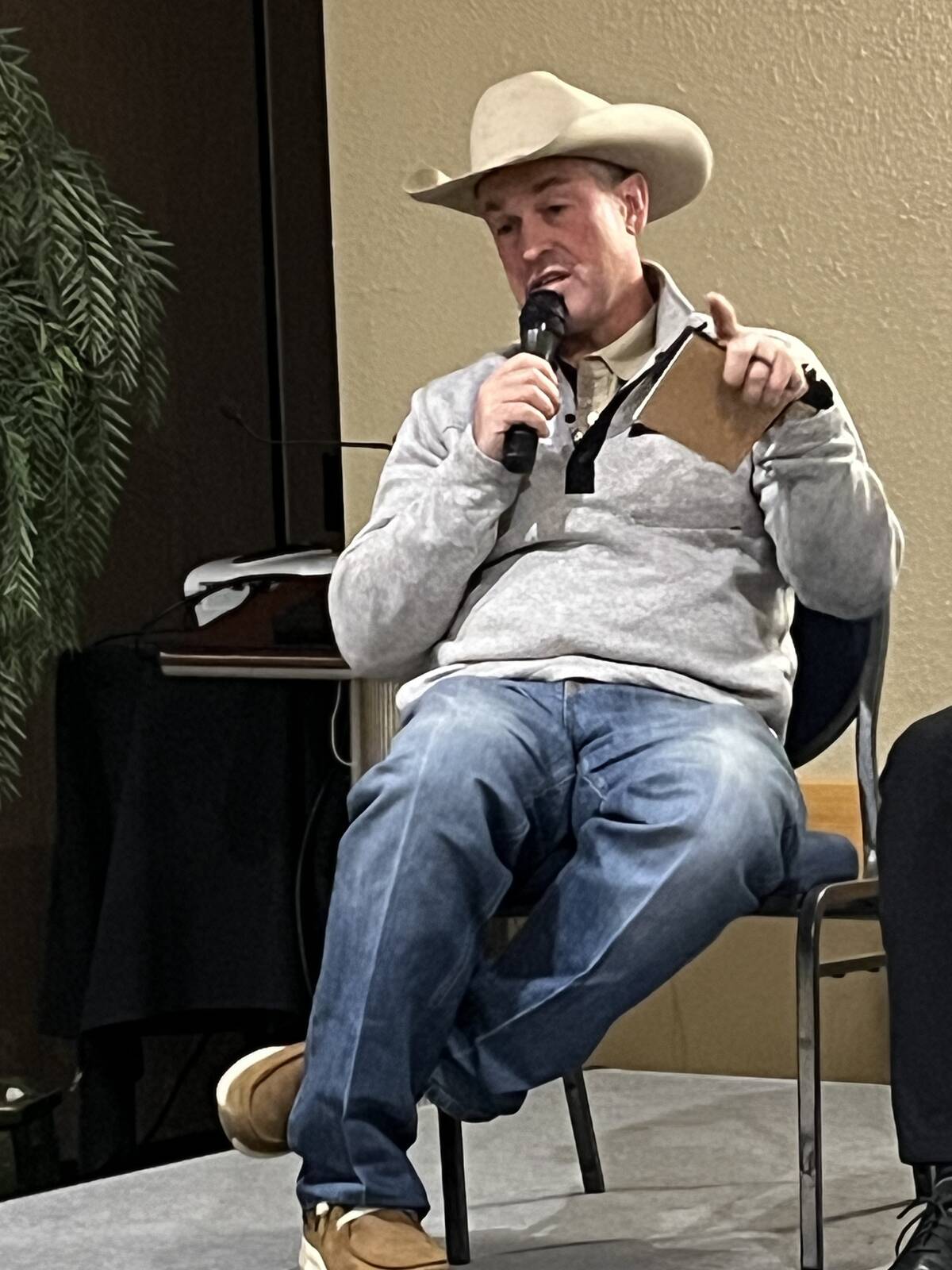 A man in a cowboy hat and blue jeans sits in a chair on a stage speaking into a microphone.