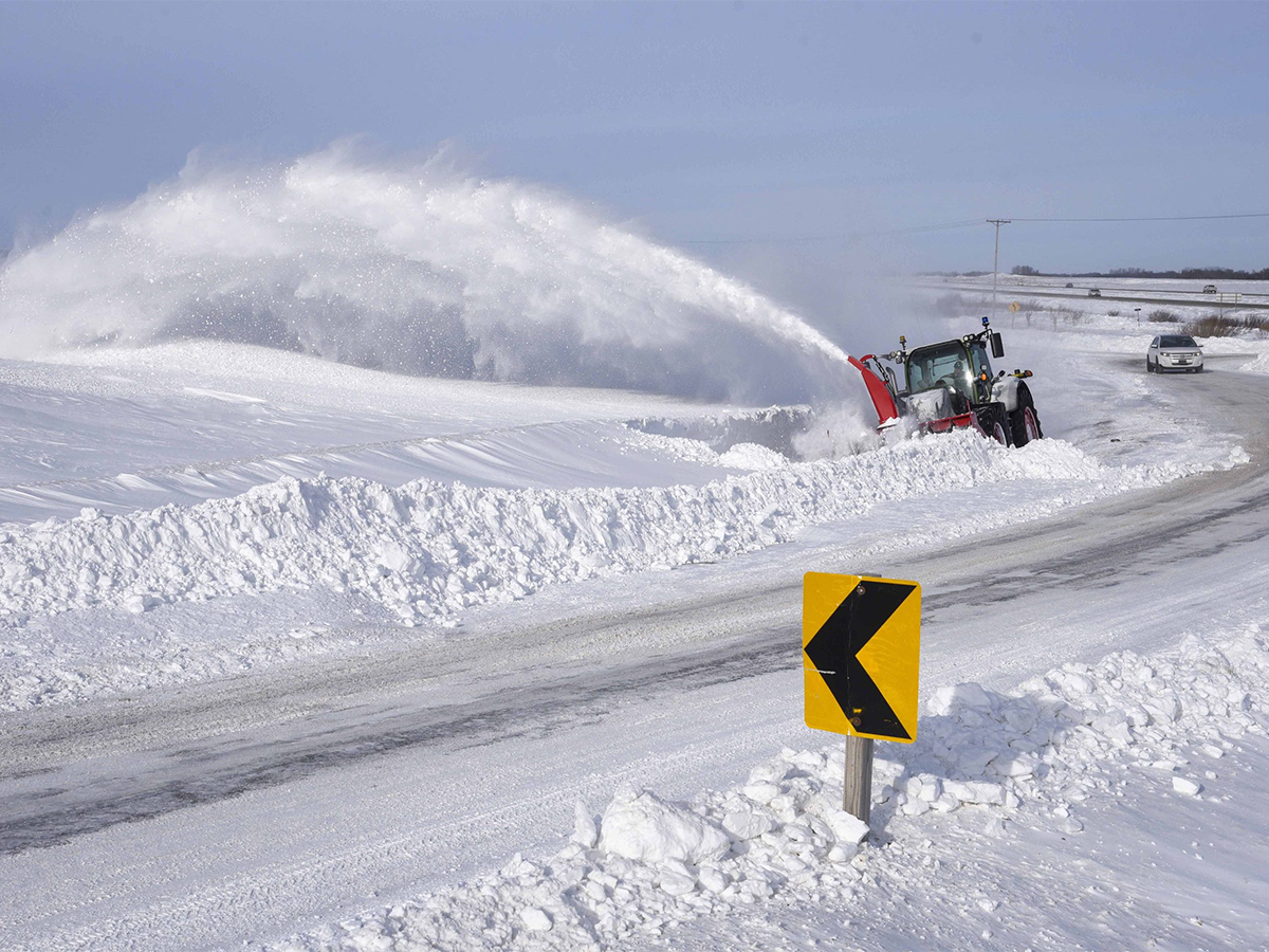 A tractor with a large, front-mounted snowblower throws snow high into the air as it clears a road.