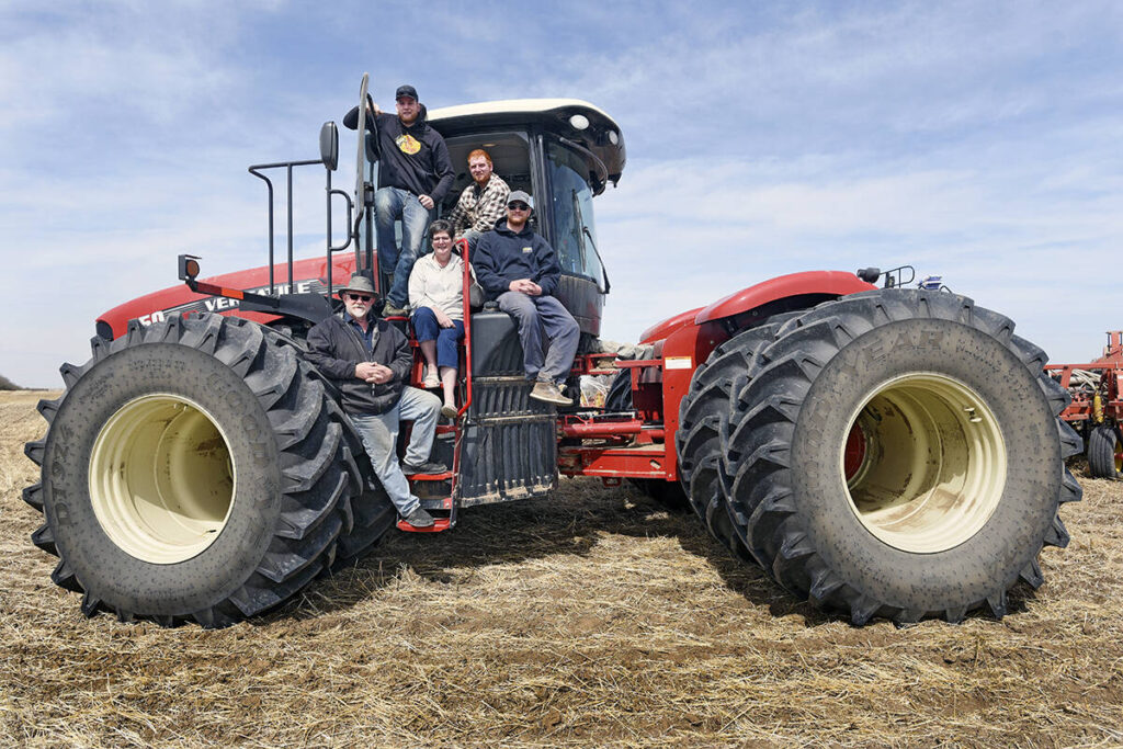 A man, his wife and their three adult sons pose for a photo on the stairs leading up to the cab of a large, Versatile brand tractor with two massive tires on each of its four corners.
