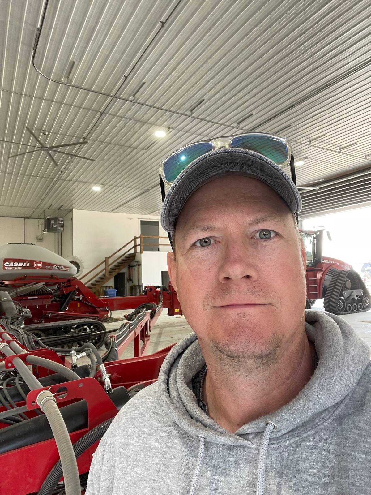 A farmer in a ball cap with his sunglasses on top of its brim stands in a large insulated equipment shop with a massive tracked tractor and air seeder rig visible behind him.