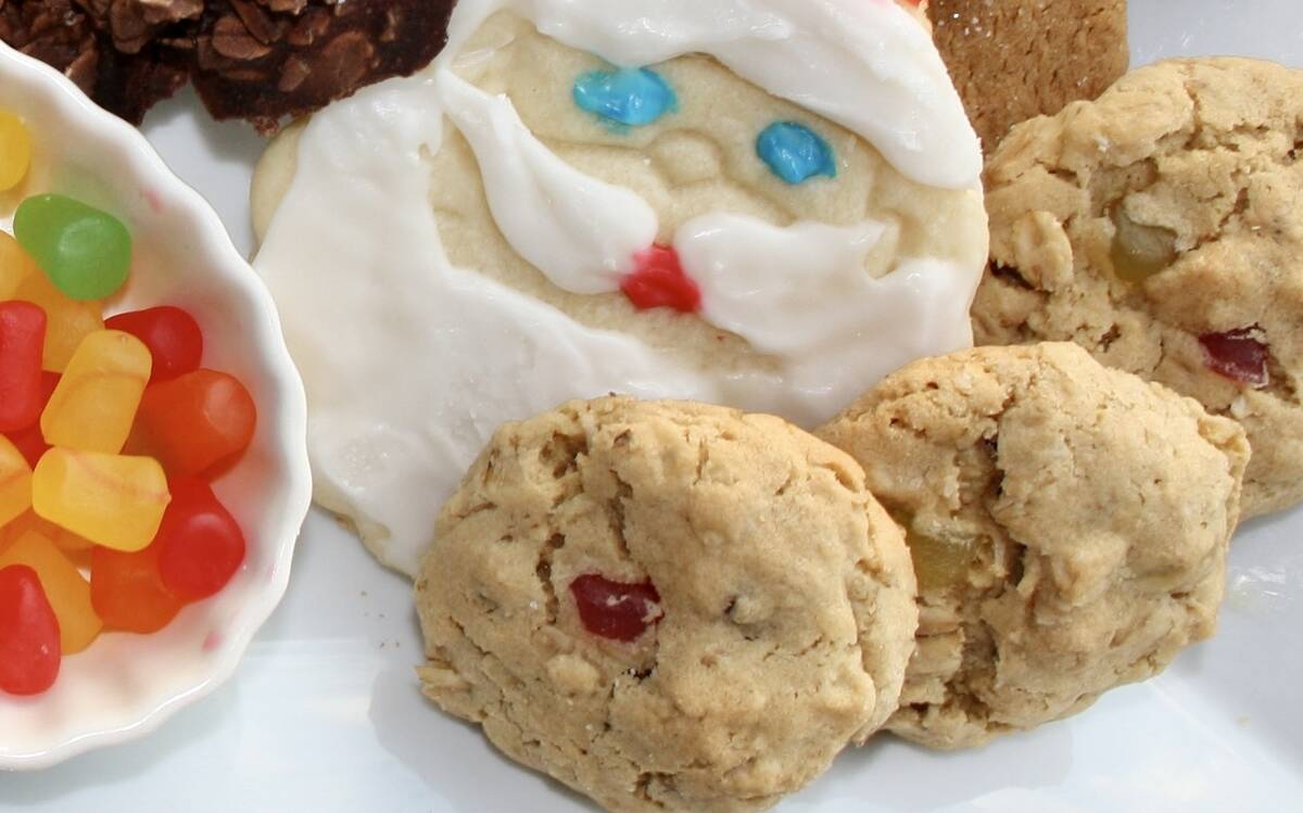 three cookies sit on a white tablecloth with a shortbread cookie with white icing on it made to look like Santa's face behind them and a bowl of gumdrops to their left.
