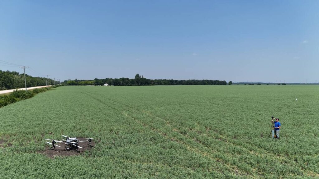 An aerial photo of a large, quad-copter style drone on a patch of dirt in an otherwise lush, green field with a man in a blue shirt standing about 10 metres to the right of the drone next to a tripod.